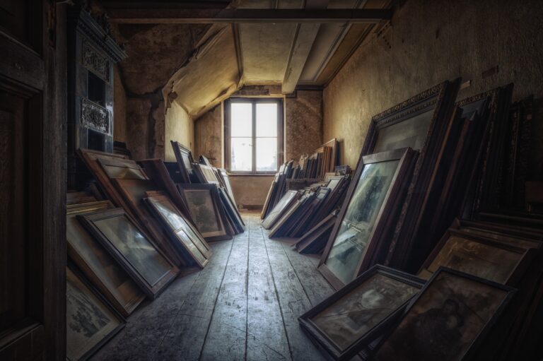 Attic with many painting and pictures stores against the walls and window straight ahead.