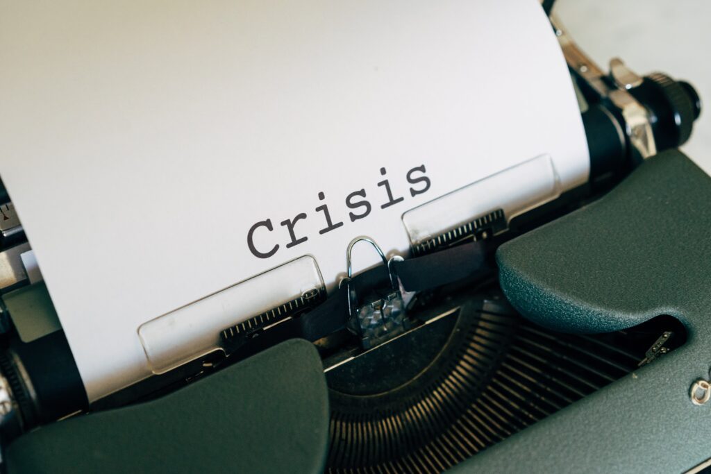 Top part of a vintage typewriter with a sheet of white paper in it. The word 'crisis' typed in black ink in the middle of the paper.