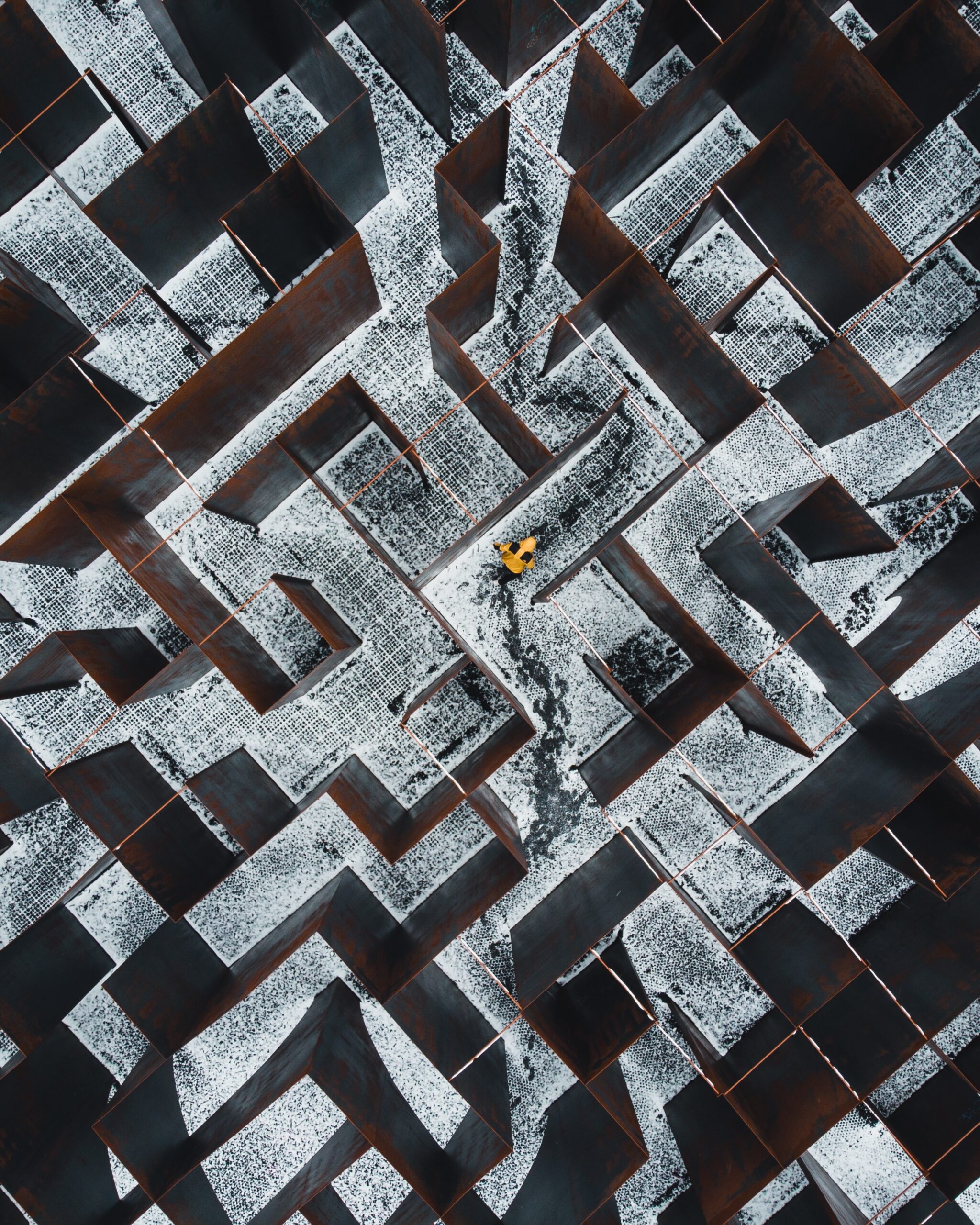 Metal maze seen from above with a figure in yellow jacket curled up. Snow on the gound