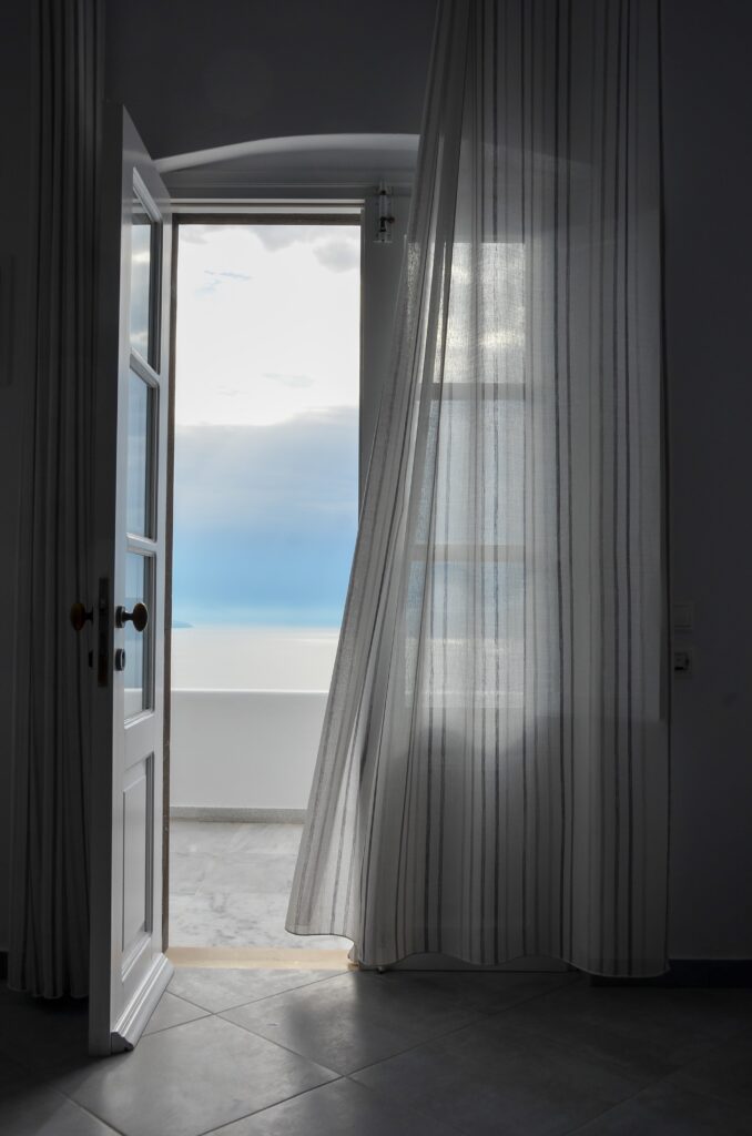 French doors onto terrace with water and mountain in blurred background. Left door open with curtain blwoing from the right in the breeze.