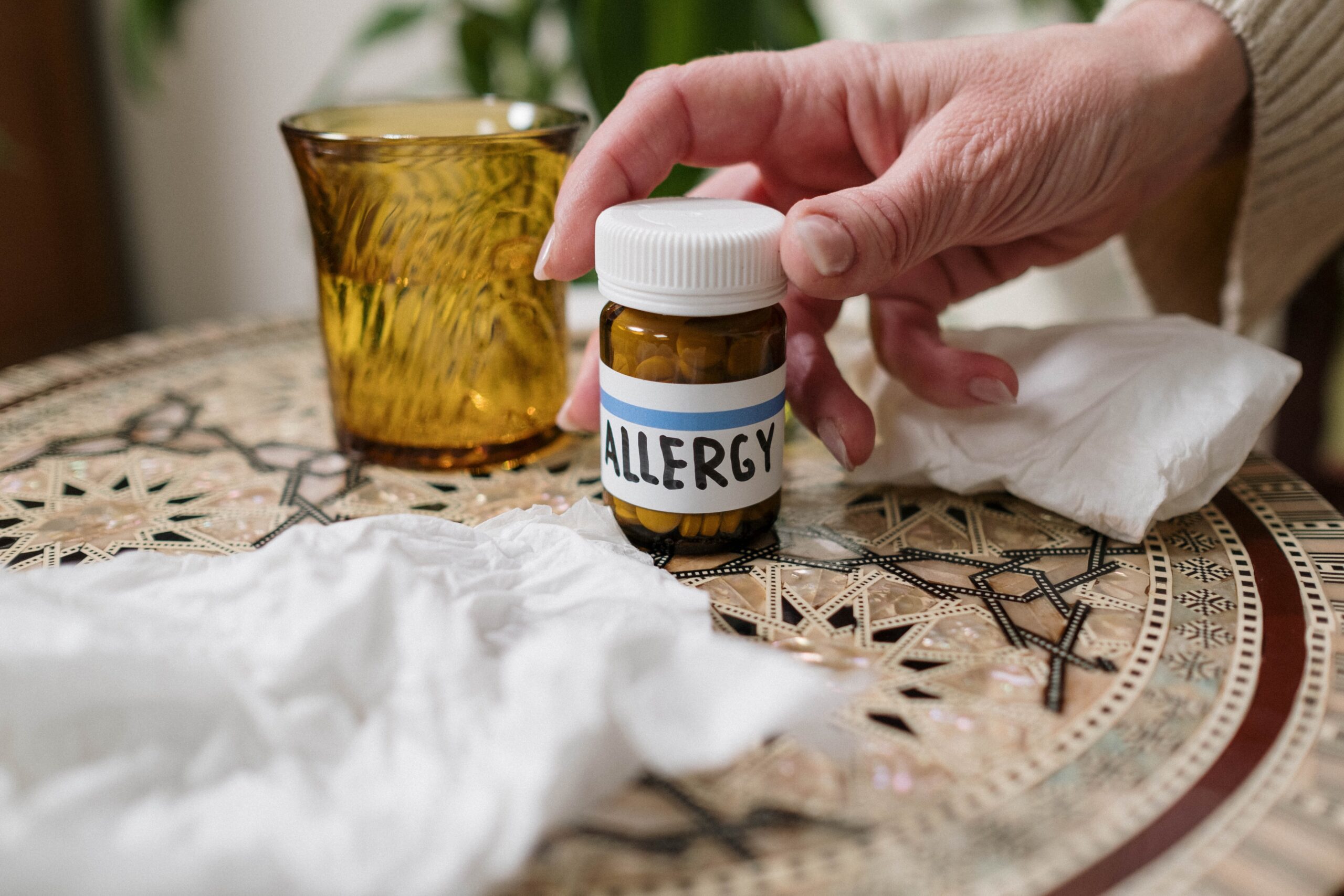 Hand reaching out to a pill bottle on the tabel with a label of 'allergy' in blue. Water glass and tissues around the bottle.