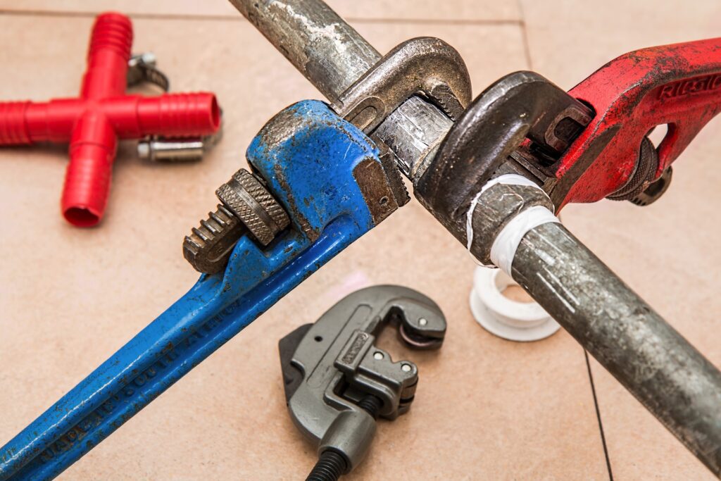 One red and one blue plumbing wrench attached to a pie with other plumbing tools on the ground in the background.