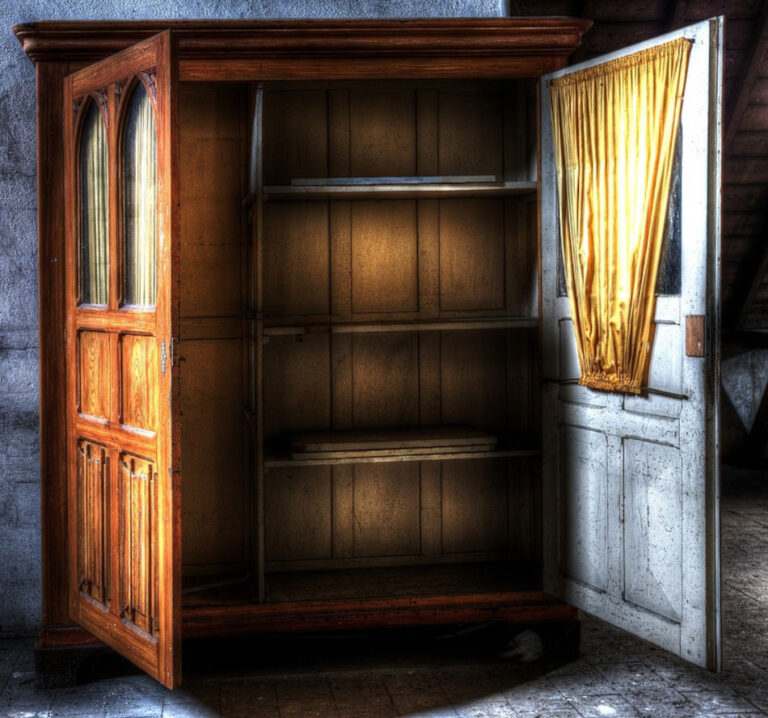 An open empty old wardrobe with double doors. Internal wooden shelves. Sets of 2 glass panels on the upper parts of both doors with yellow curtains on the inside.